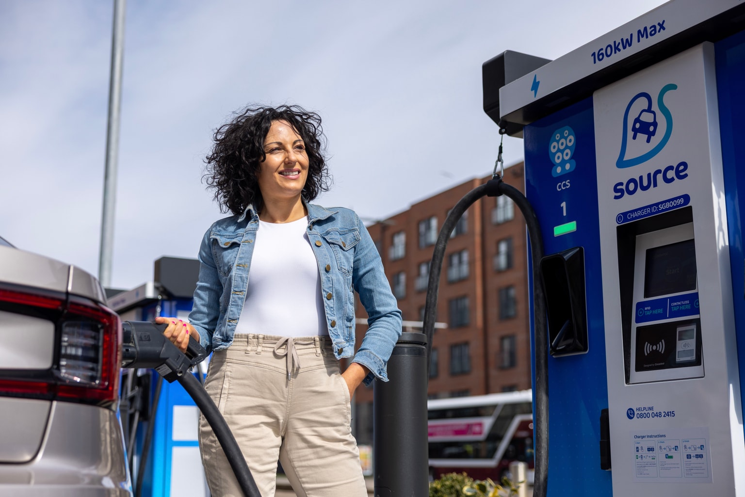 a woman charging her EV at a dark blue, fast Source charger with a modern building and cloudy blue skies in the background.