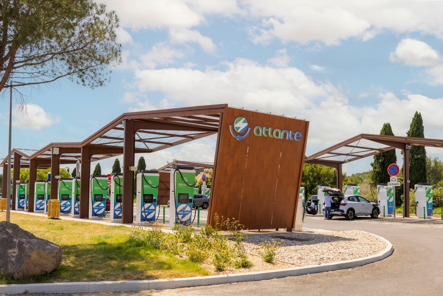 Una grande struttura in legno copre le colonnine di ricarica per veicoli elettrici Atlante, immerse nel verde e sotto un cielo azzurro e nuvoloso. In lontananza, una persona prende qualcosa dal bagagliaio della propria auto.