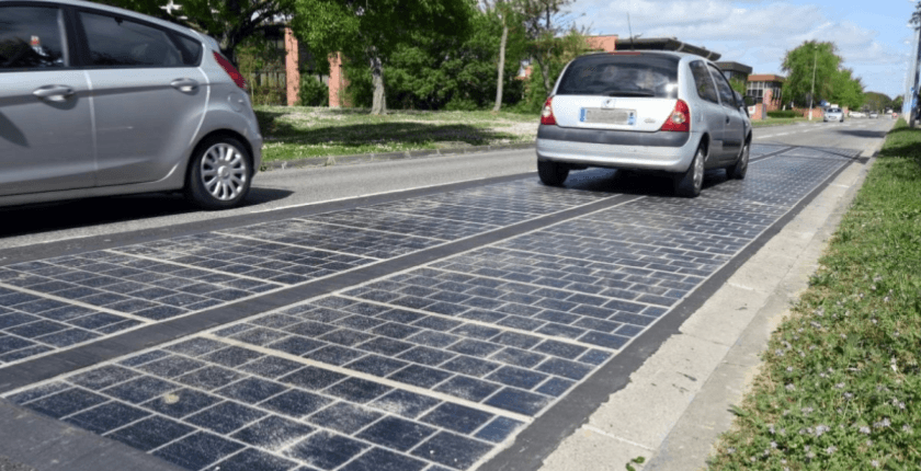Cars driving over a road with integrated solar panels on a sunny day, surrounded by greenery and buildings.