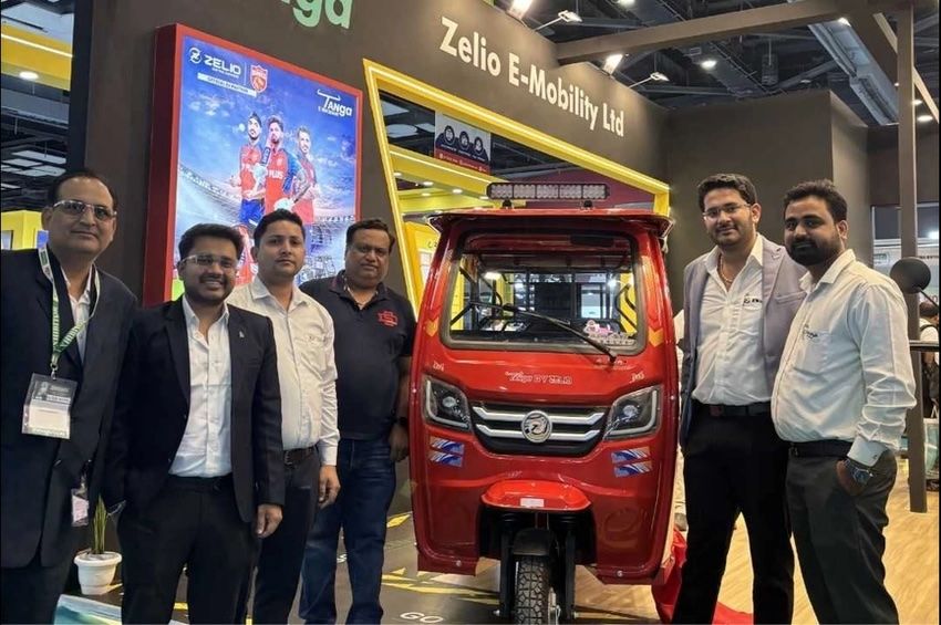 Group of people standing beside a red electric rickshaw at the Zelio E-Mobility Ltd booth, with a promotional poster in the background.