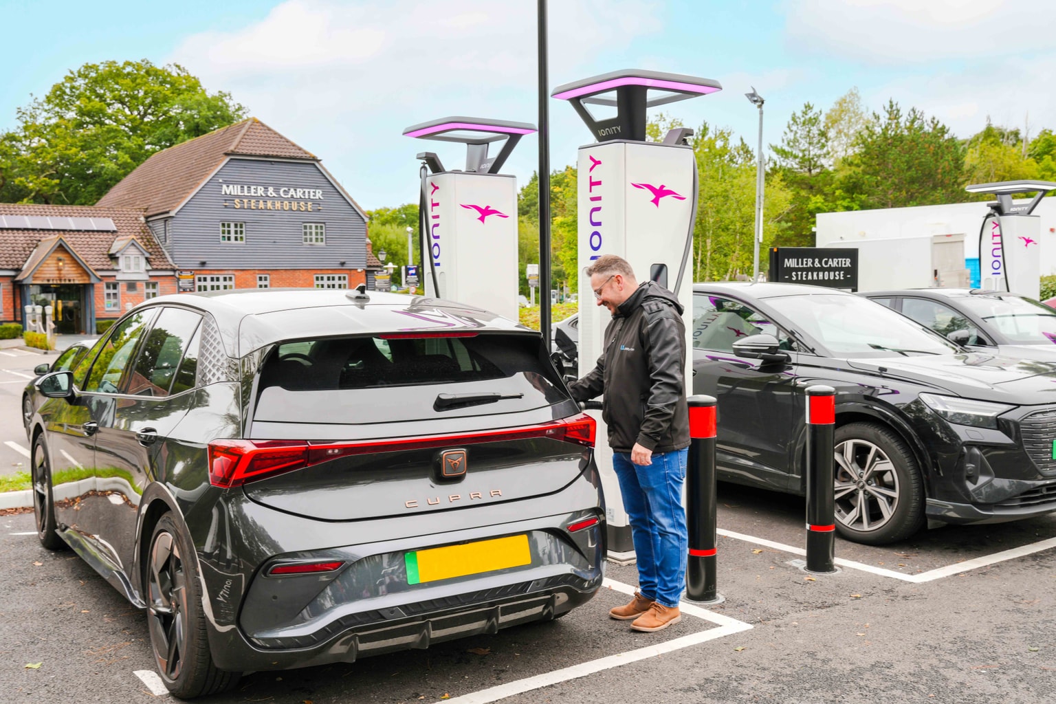 Uomo che ricarica un'auto elettrica nera presso una stazione di ricarica in un parcheggio vicino a un edificio con la scritta "Miller & Carter".