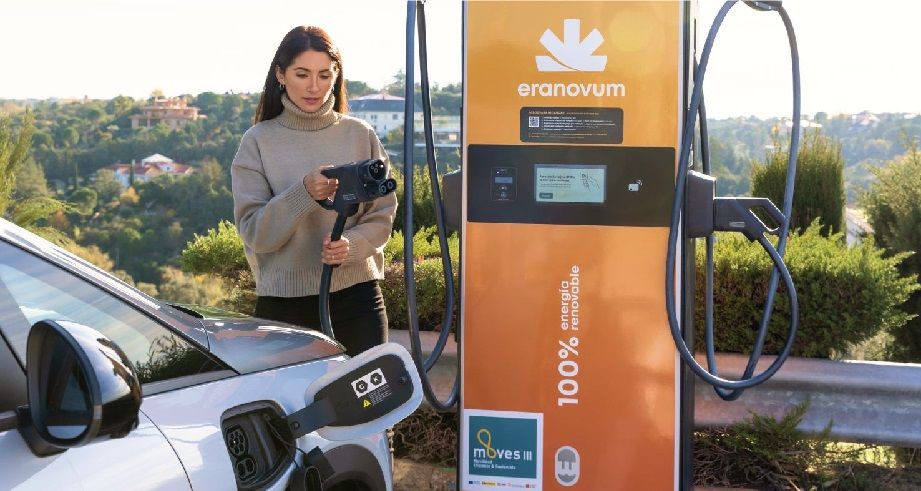 Una mujer carga un coche eléctrico en una estación de carga Eranovum de color naranja al aire libre, rodeada de vegetación y edificios distantes.