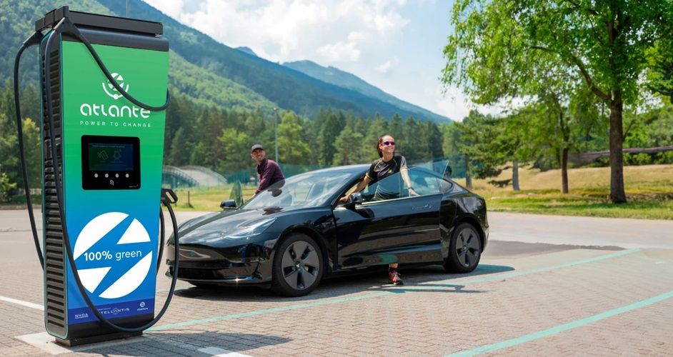 Un'auto elettrica in carica presso una stazione Atlante con etichetta "100% verde", situata in una pittoresca zona montana, con due persone vicino al veicolo.