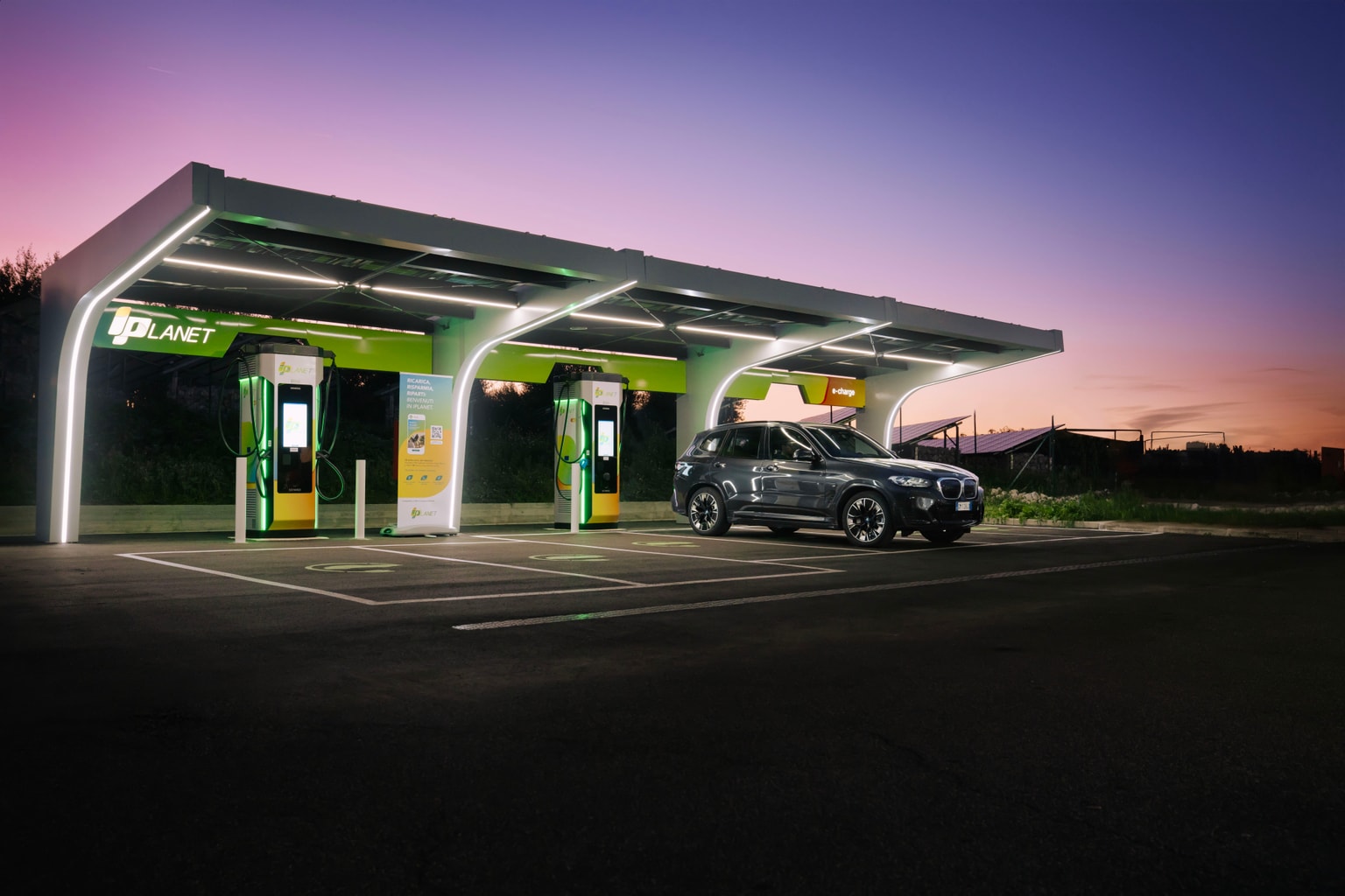 Un'auto si ricarica presso una stazione elettrica iPlanet illuminata sotto un cielo crepuscolare, con accenti verdi e un design moderno.