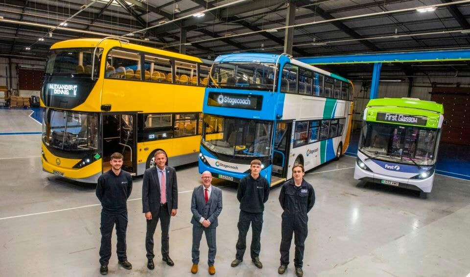 Quattro persone sono in piedi davanti a tre autobus a due piani in un magazzino, ognuno dipinto con colori e design diversi.