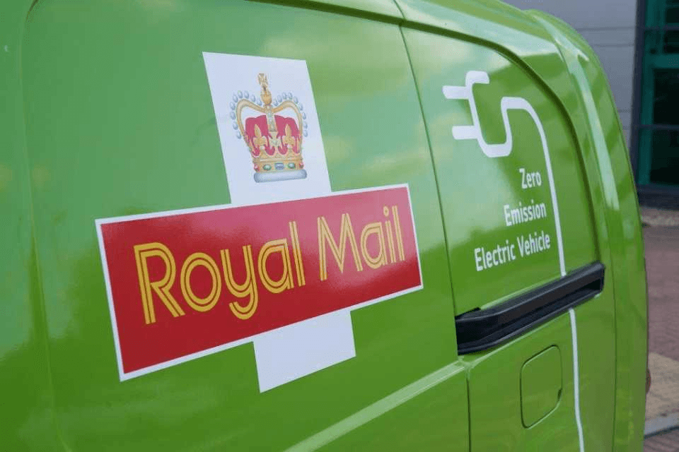 Furgone verde della Royal Mail con il logo di una corona e la scritta "Zero Emission Electric Vehicle" sulla fiancata, parcheggiato vicino a un edificio.