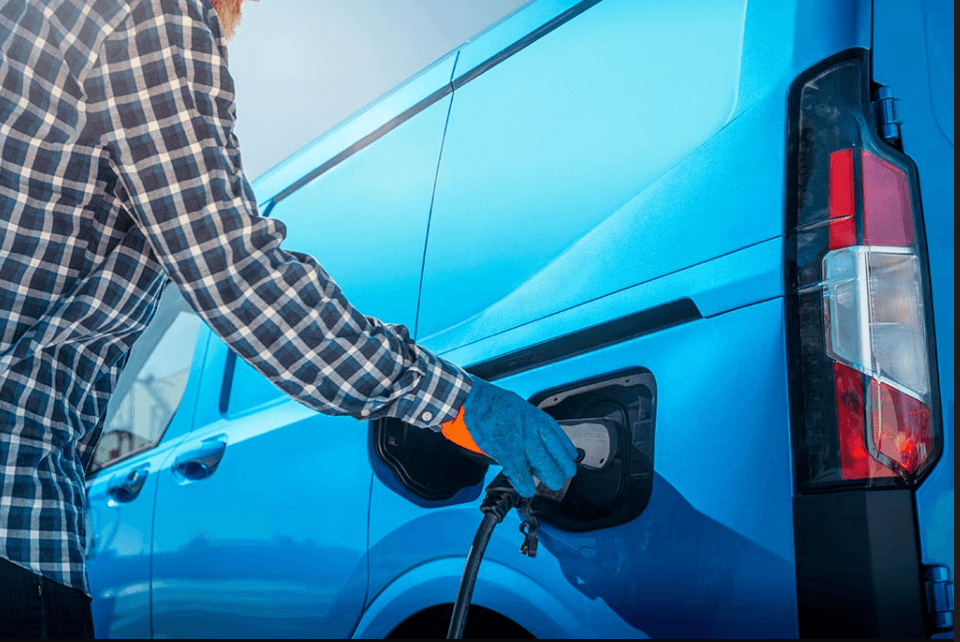 Person in a plaid shirt and blue gloves plugs a charger into a bright blue electric van.