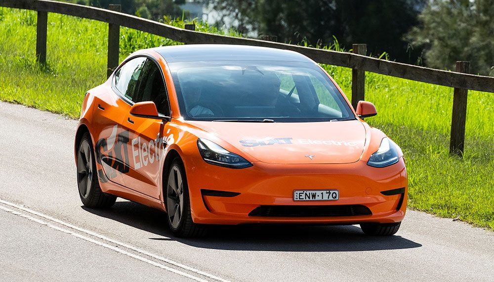 Coche eléctrico naranja con la marca «Sixt Electric» circulando por una carretera rural, rodeado de vegetación y una valla de madera.