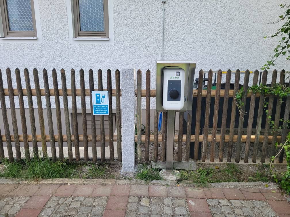 Cocharge-Ladestation für Elektrofahrzeuge an einem Holzzaun neben einem Schild mit einem Ladesymbol vor einer weißen Gebäudewand.