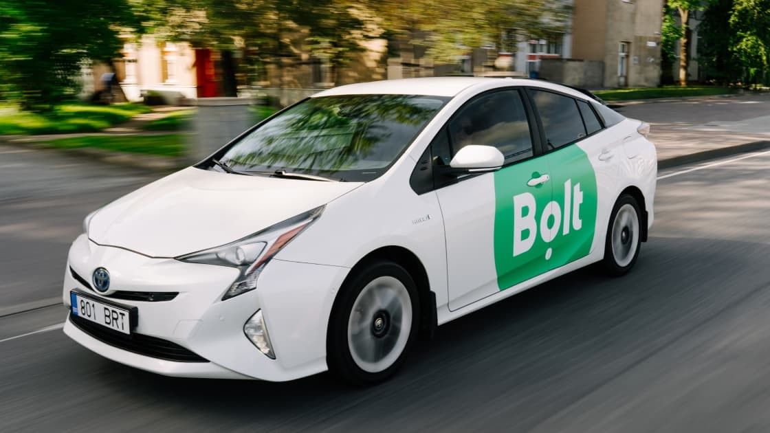 A white Toyota Prius with a green "Bolt" logo on the side is driving on a city street.