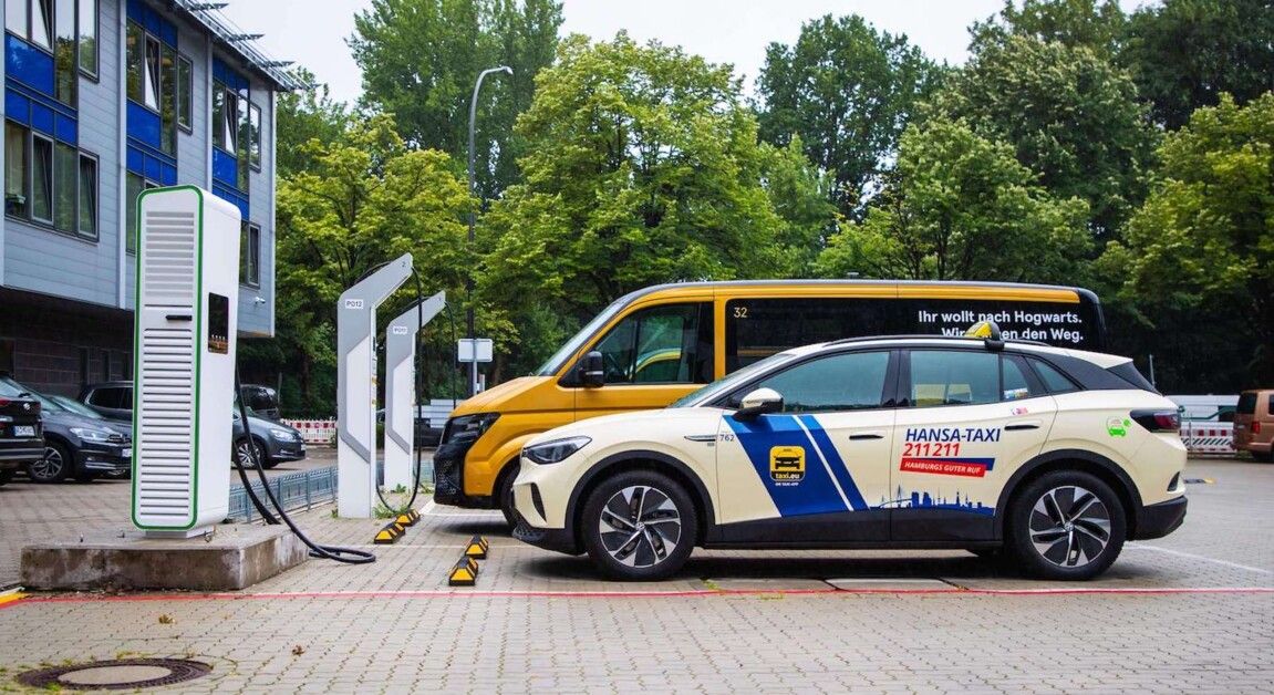 Elektrotaxi mit „Hansa-Taxi”-Logo an der Seite und gelber Lieferwagen, die an einer Station in einem von Bäumen gesäumten Parkplatz mit Gebäuden in der Nähe aufgeladen werden.