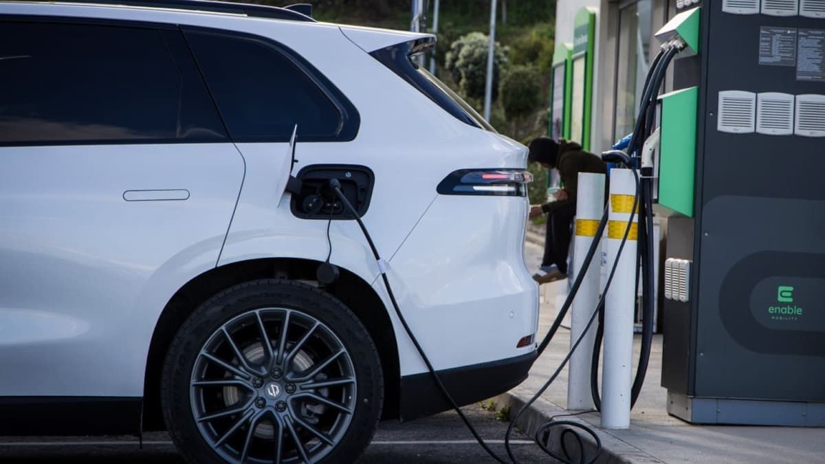 SUV elettrico bianco che si ricarica in una stazione, con un cavo collegato alla parte posteriore del veicolo. Si vede una persona sullo sfondo.