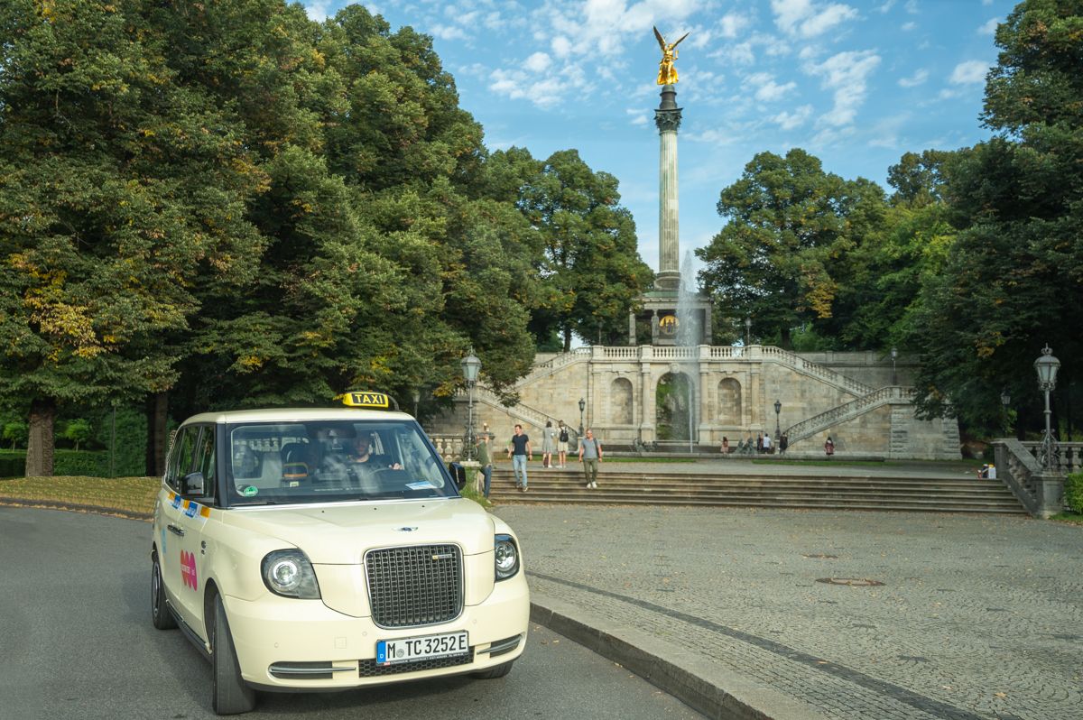 Ein weißes Londoner Taxi fährt am Friedensengel-Denkmal in München vorbei, umgeben von üppigen Bäumen und einem strahlend blauen Himmel.