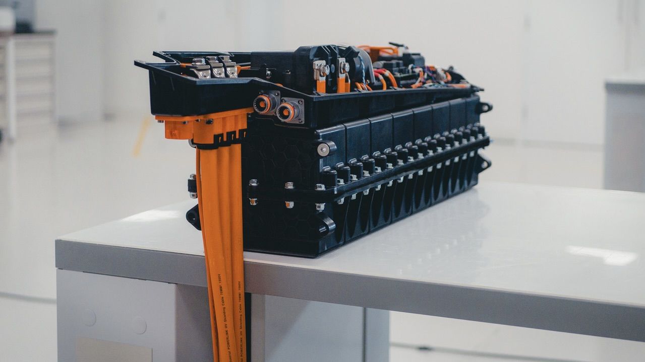 Primer plano de un paquete de baterías de un vehículo eléctrico con cables naranjas sobre una mesa blanca en una habitación luminosa.