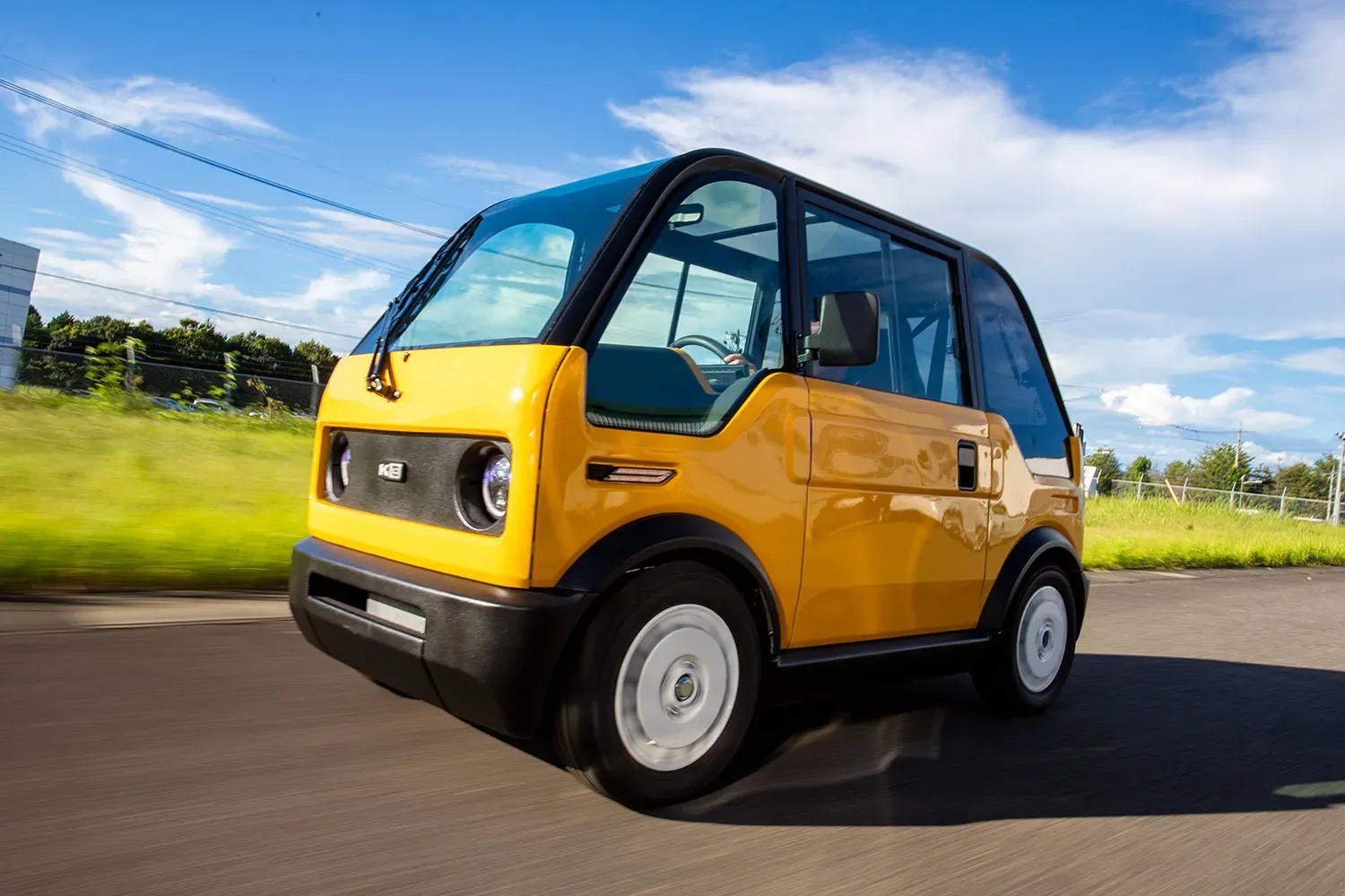 Un vehículo eléctrico compacto, de color amarillo y con grandes ventanas, circula por una carretera junto a un césped verde bajo un cielo azul con nubes.