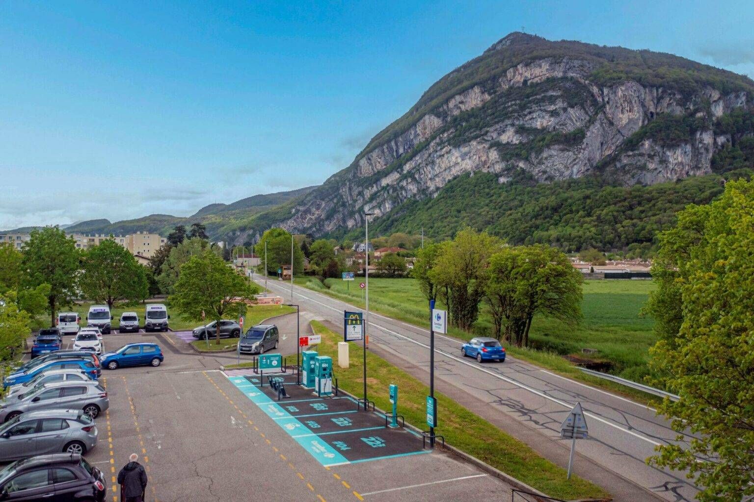 Un parking équipé de bornes de recharge pour véhicules électriques, entouré d'arbres et d'un paysage montagneux sous un ciel clair.