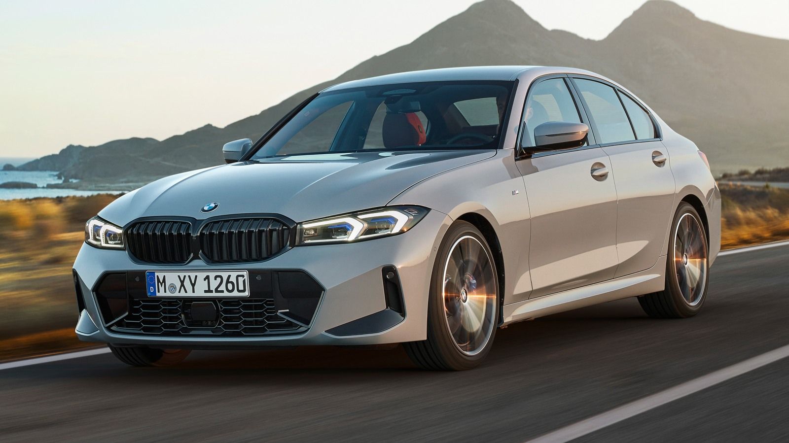 Ein grauer BMW 3er fährt auf einer Straße entlang, im Hintergrund Berge, Gras und das Meer.