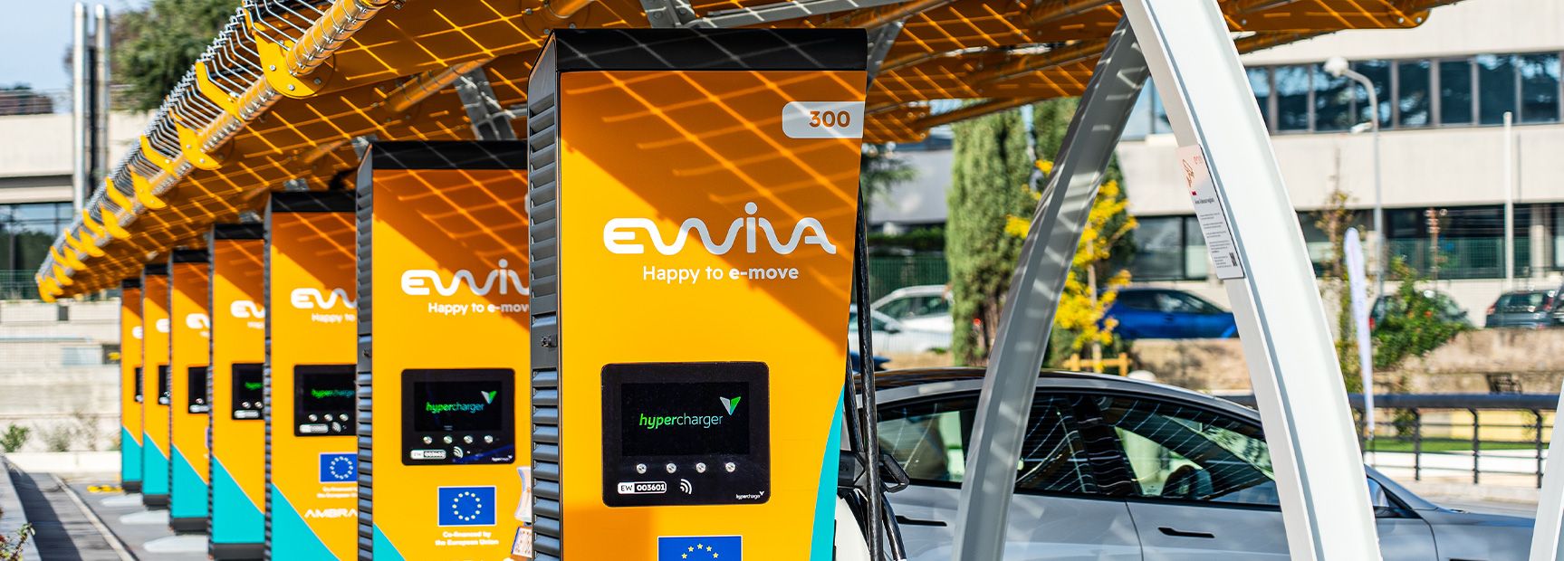 Diverse stazioni di ricarica per veicoli elettrici Ewiva con pannelli arancioni e un'auto parcheggiata sotto una tettoia solare.