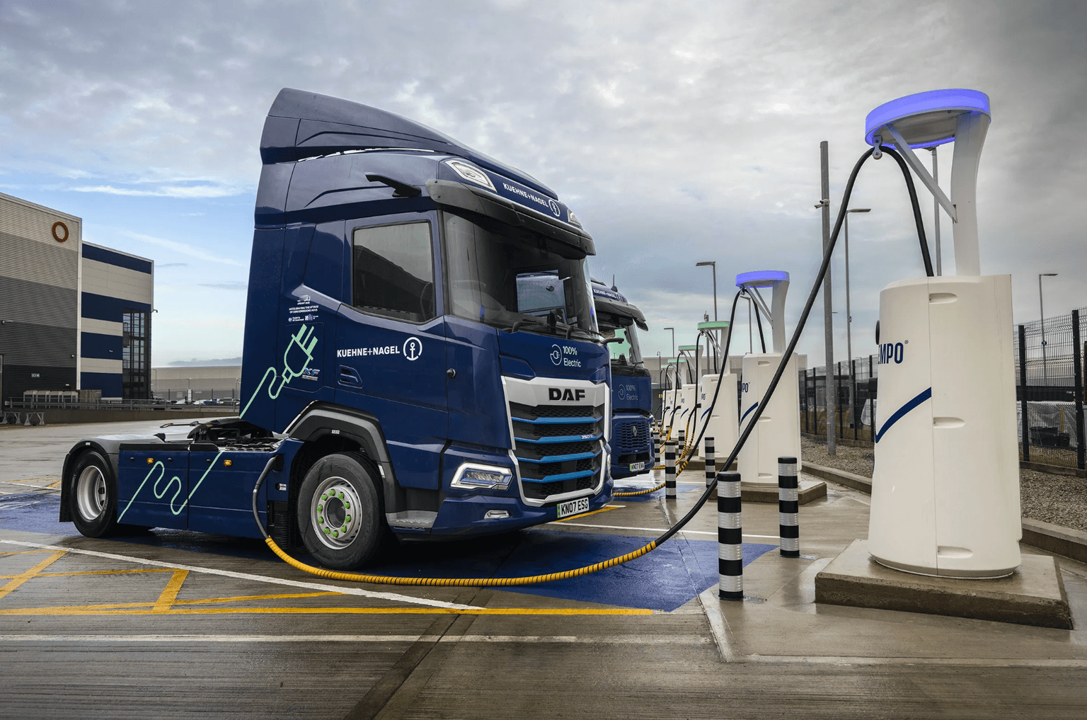 Electric truck at a charging station, connected via a yellow cable. Overcast sky, industrial background with warehouse and poles.