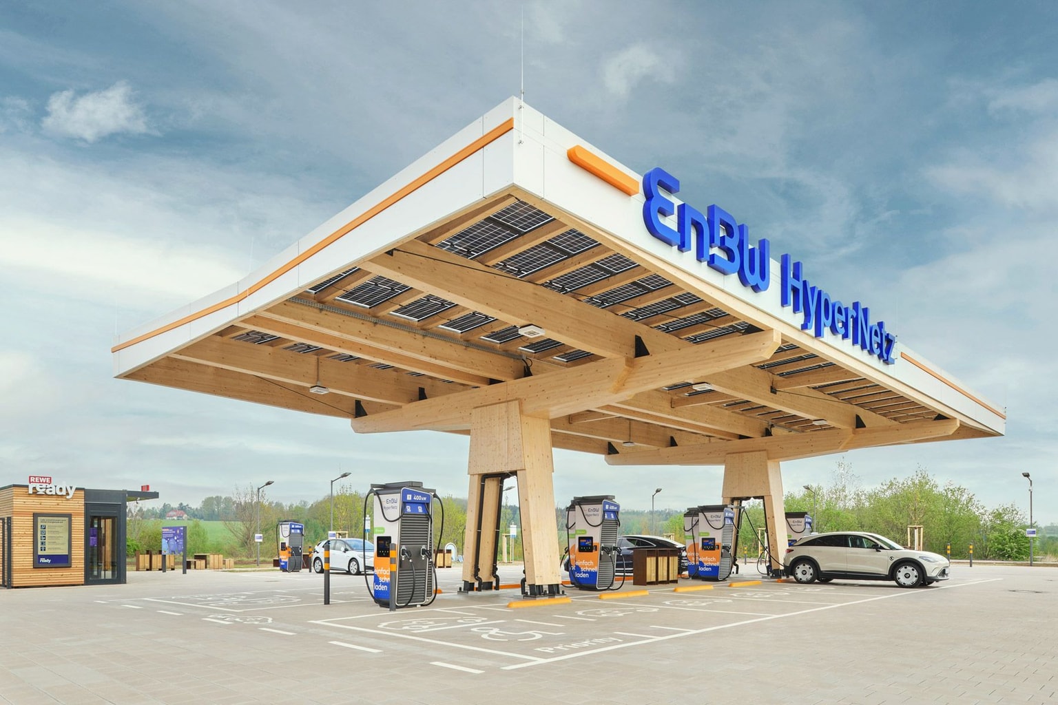 Ein großes, blaues EnBW HyperNetz-Logo auf einem großen Holzdach, das vier Schnellladegeräte für Elektrofahrzeuge und ein Elektrofahrzeug abdeckt, mit weiteren Ladegeräten und Grünpflanzen im Hintergrund, unter einem blauen Himmel mit vielen dünnen weißen Wolken.