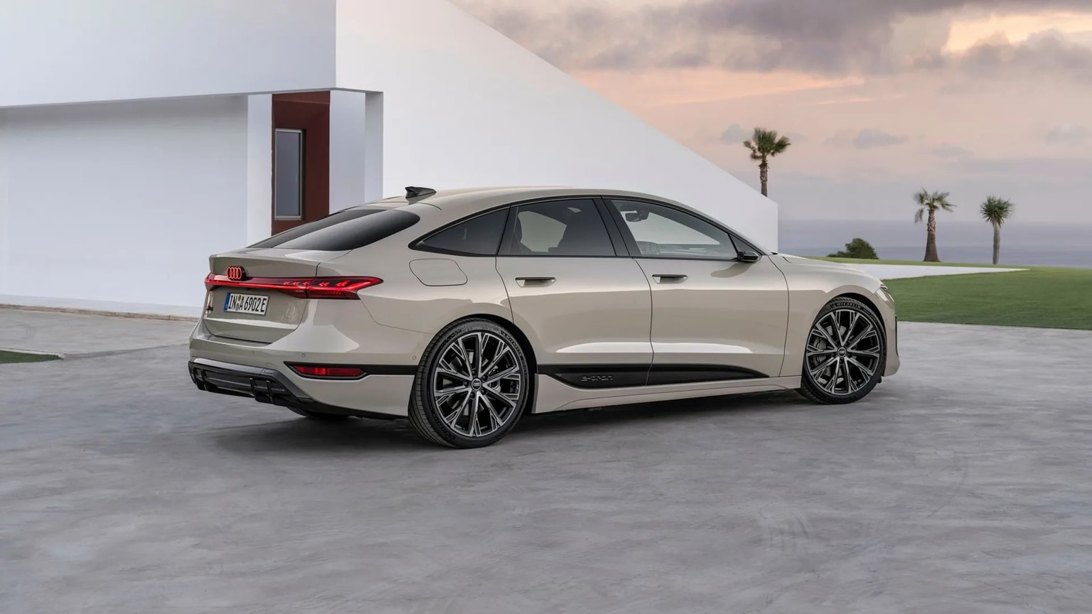 Ein Foto eines Audi A6 e-tron, der auf einer grauen Betonauffahrt vor einem modernen Gebäude mit Bäumen und dem Meer im Hintergrund geparkt ist.