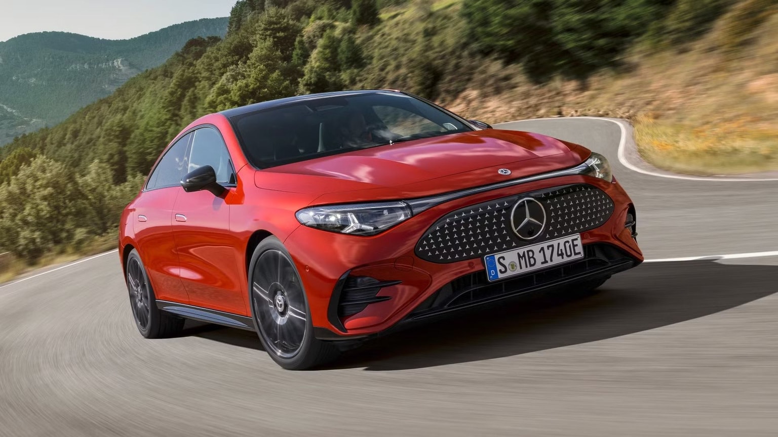 Un sedán Mercedes-Benz rojo circulando por una carretera sinuosa con árboles y colinas al fondo.