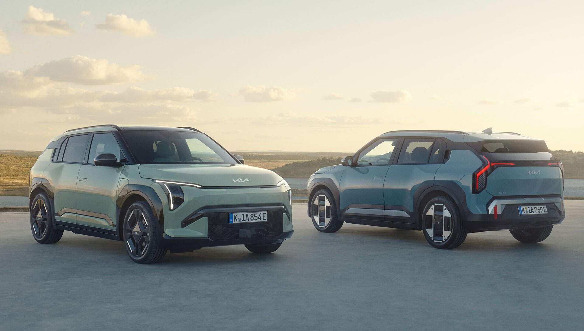 Dos SUV KIA EV-3 en un área de estacionamiento, uno mirando hacia adelante y el otro hacia atrás, bajo un cielo despejado con un paisaje escénico de fondo.