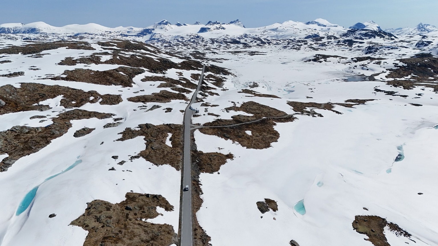 Aerial view of a winding road through a snowy, mountainous landscape with patches of brown terrain and distant snow-capped peaks under a clear sky.