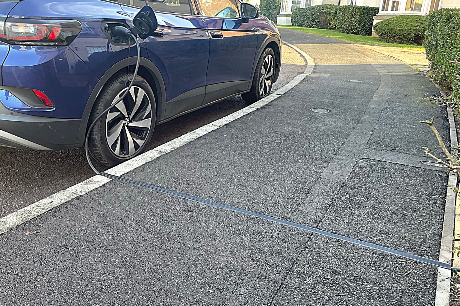 Coche eléctrico azul, aparcado en una entrada, cargándose con un cable que se extiende por la acera.