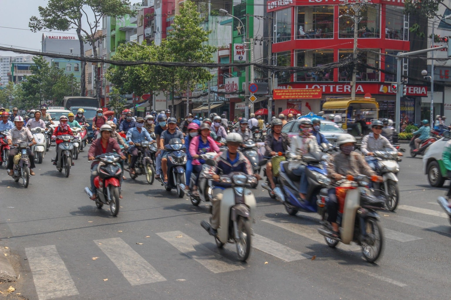 Una concurrida calle urbana con numerosos motociclistas con casco, cruzando un cruce, rodeados de edificios y tráfico.