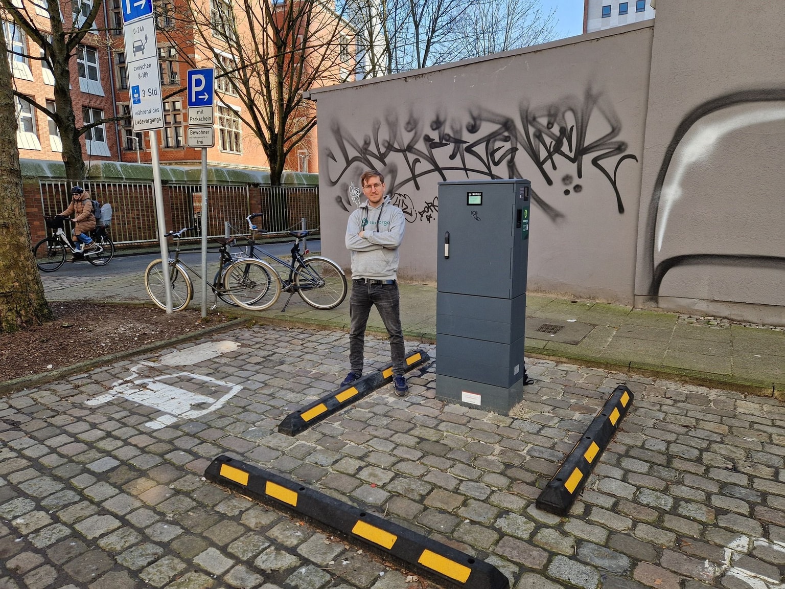 Mann steht auf einer Kopfsteinpflasterstraße in der Nähe einer Cocharge-Ladestation mit Graffiti an der Wand und in der Nähe geparkten Fahrrädern.