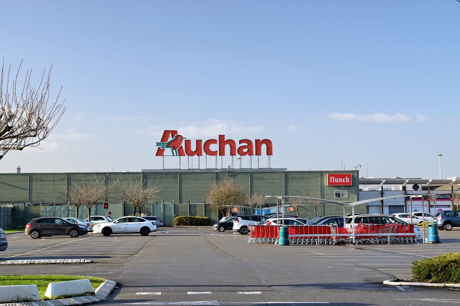 Parking devant un supermarché Auchan par temps clair, avec des voitures garées et une zone de retour des caddies visible.
