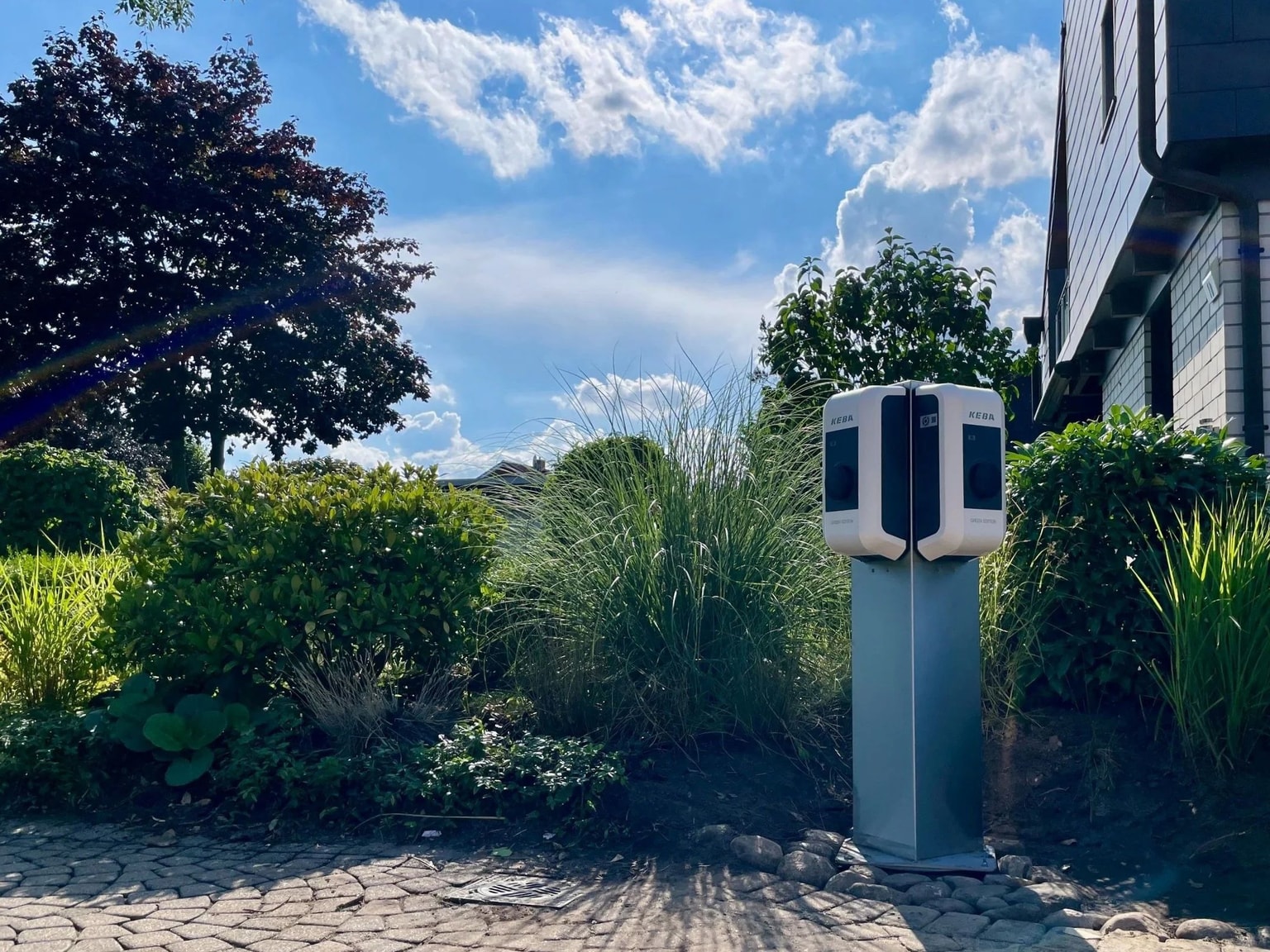 Cocharge-Ladestation für Elektrofahrzeuge, umgeben von üppigem Grün und unter einem teilweise bewölkten blauen Himmel in der Nähe eines Gebäudes.