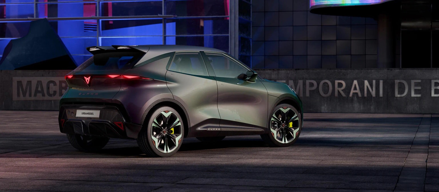 Ein Cupra Reval, geparkt auf einer Straße mit dunklem Hintergrund, modernen Gebäuden und einer Werbung.