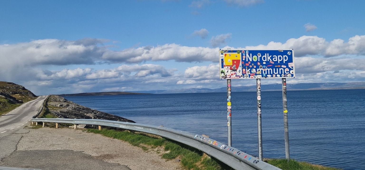 A coastal road leads to a colorful sign reading "Nordkapp Kommune" by a calm sea under a blue sky with scattered clouds.