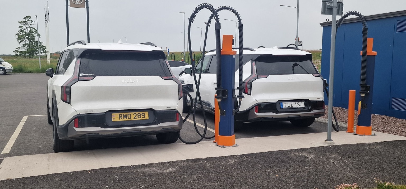 Two white electric Kia SUVs charging at a station, with charging cables connected. Overcast sky and a blue building in the background.