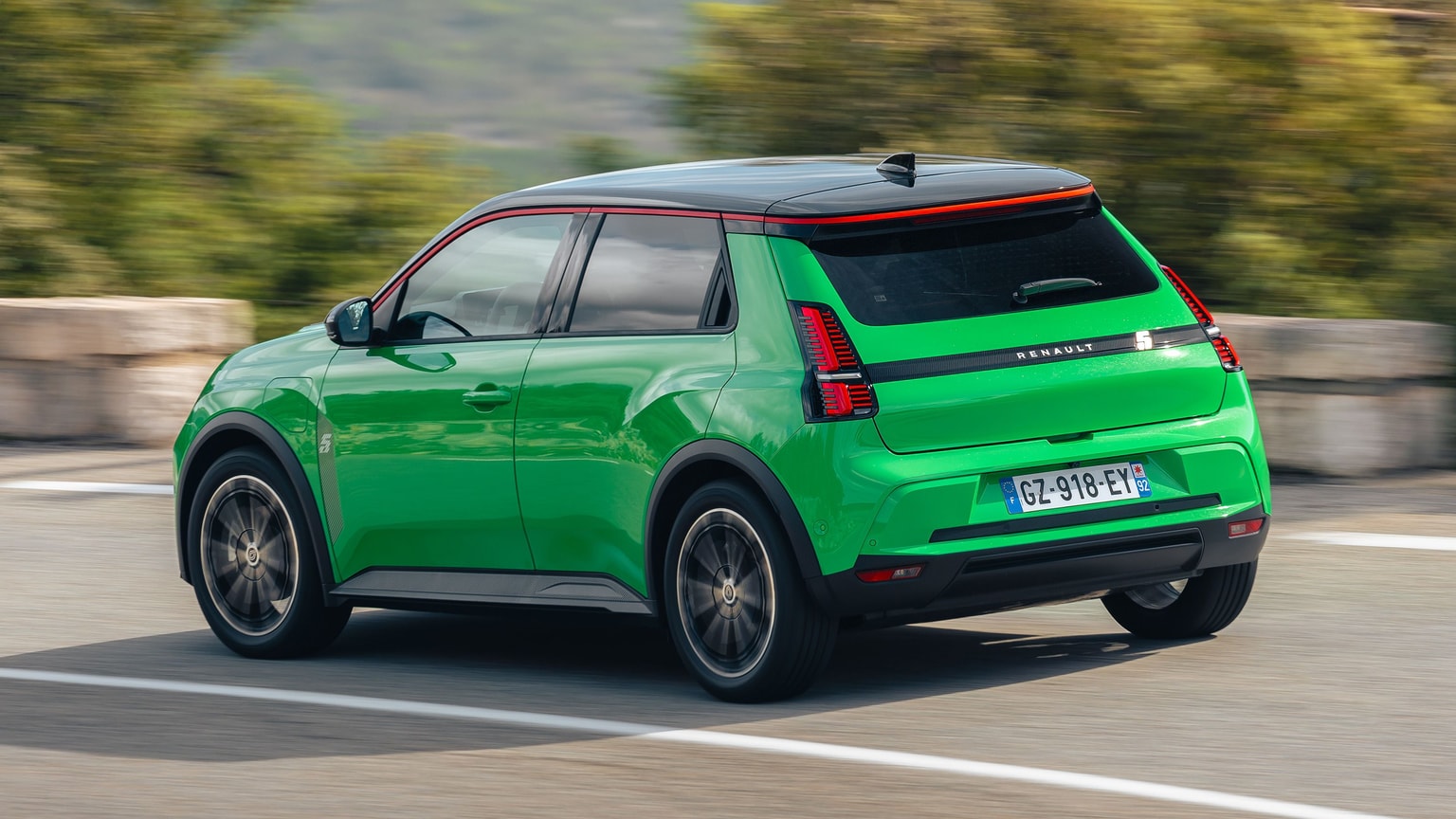 Una vivace auto compatta verde con il tetto nero sfreccia su una strada, circondata dal verde e da immagini sfocate, mettendo in risalto il suo design elegante.