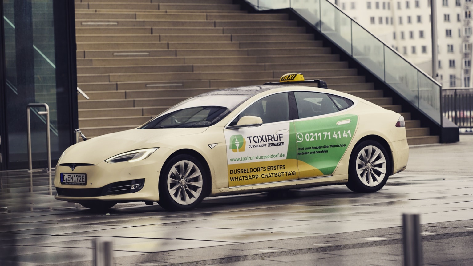 Ein weißes Tesla-Taxi mit grün-gelbem „TaxiRuf”-Branding steht auf einer nassen Straße in der Stadt in der Nähe moderner Gebäude und Treppen.