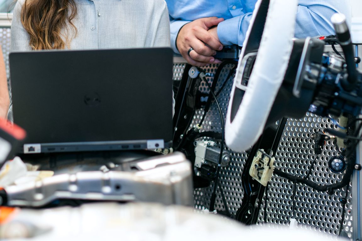 Dos personas trabajan en una computadora portátil cerca de equipos automotrices, incluido un volante, en un entorno técnico.