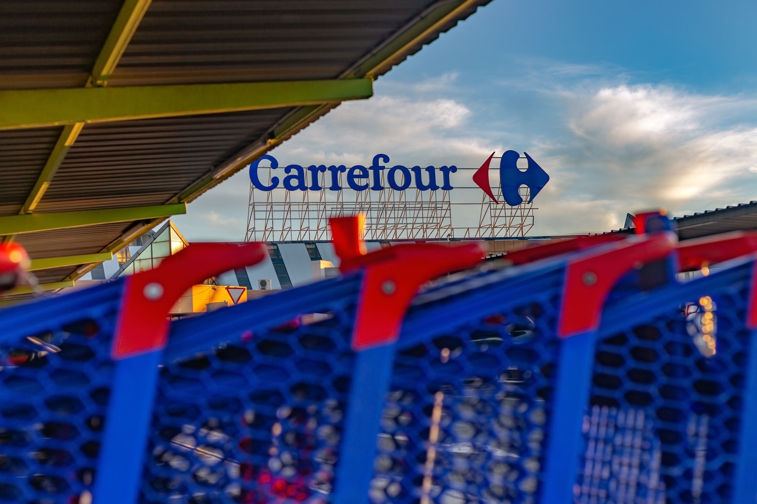 Une enseigne du supermarché Carrefour au-dessus d'une rangée de caddies bleus à poignées rouges, avec un ciel bleu et des nuages blancs en arrière-plan.
