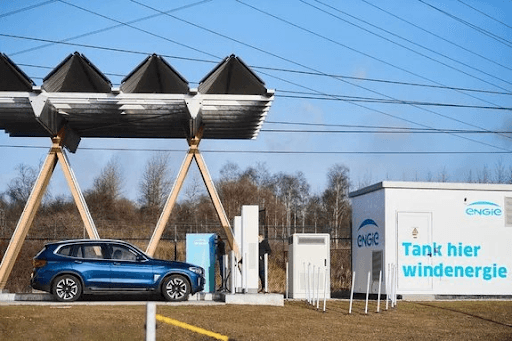 Coche azul en una estación de recarga de energía eólica bajo paneles solares, con un cartel que dice «Tank hier windenergie» (Recarga aquí energía eólica) y líneas eléctricas al fondo.