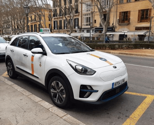Taxi blanco aparcado en una calle de la ciudad, con rayas naranjas y un letrero de taxi en el techo, edificios y árboles al fondo.