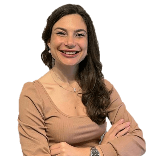 Greta sorridente, addetta al marketing di iPlanet, con lunghi capelli castani, indossa un top beige, una collana e un orologio, con le braccia incrociate, su uno sfondo bianco.