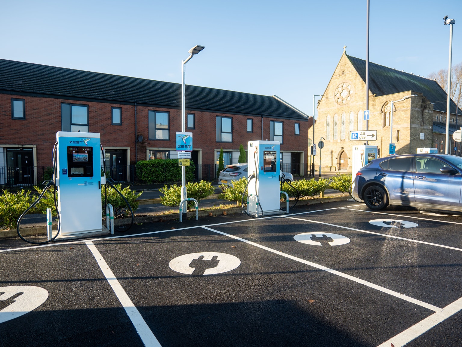 EV charging station bays with Zest chargers and painted plug symbols, blue car parked, brick row houses and church in the background.