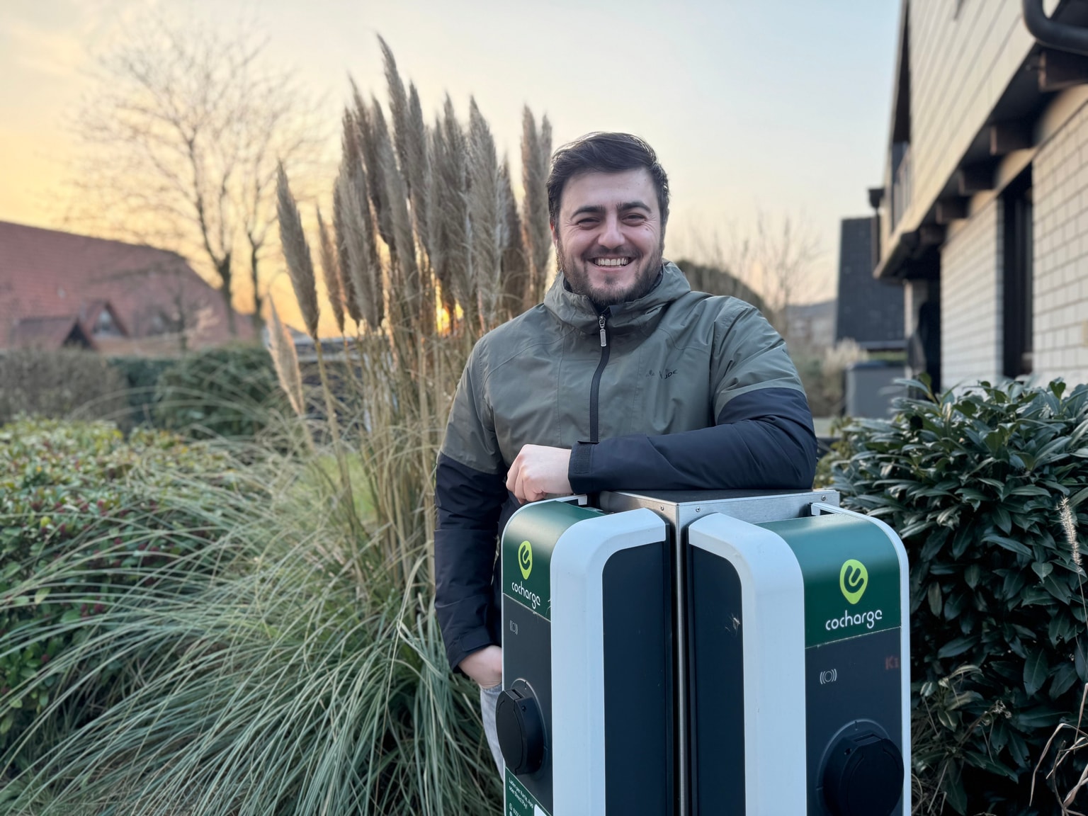 Eine Person in einer Jacke lehnt lächelnd an zwei Ladestationen, im Hintergrund hohes Gras und ein Haus bei Sonnenuntergang.