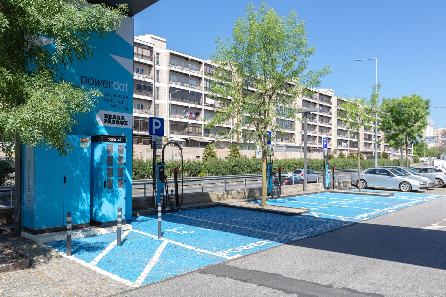 Estación de carga de vehículos eléctricos con plazas de aparcamiento azules, rodeada de árboles y adyacente a un edificio residencial de varios pisos.