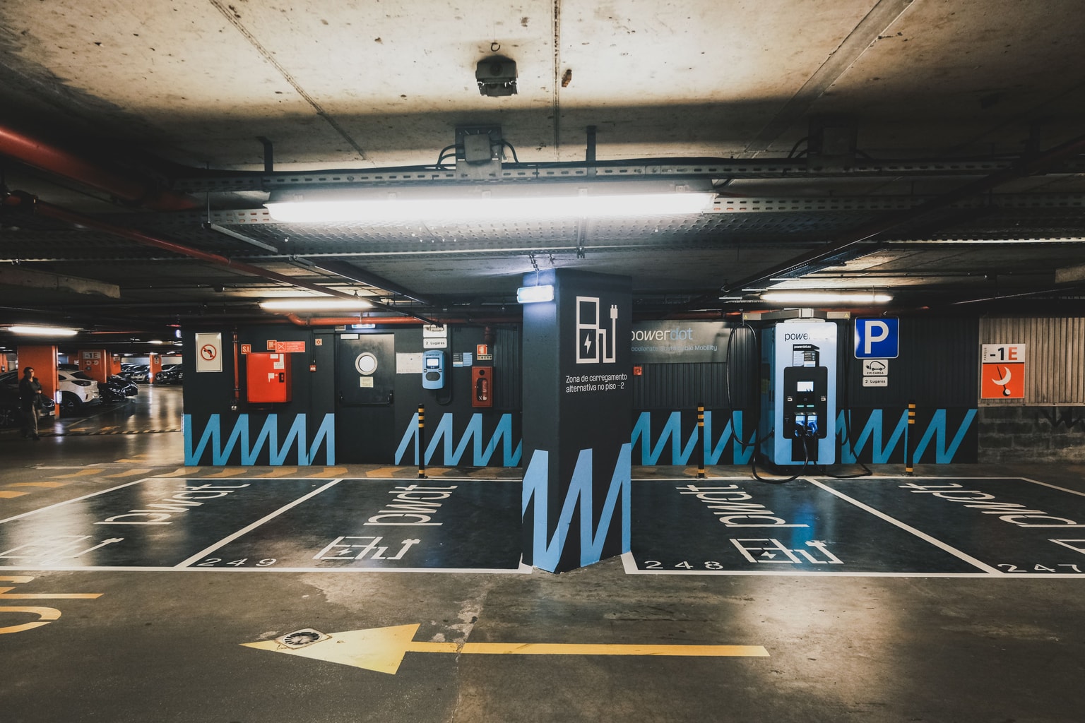Estacionamiento subterráneo con estaciones de carga para vehículos eléctricos Powerdot designadas, marcadas con líneas azules en zigzag y unidades de carga en la pared.