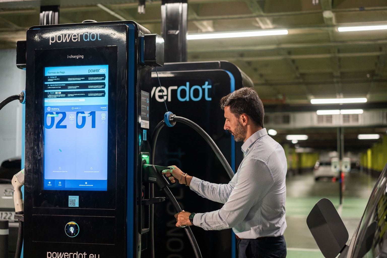 Un hombre carga un vehículo eléctrico en una estación de carga Powerdot en un estacionamiento. La estación muestra información de carga.