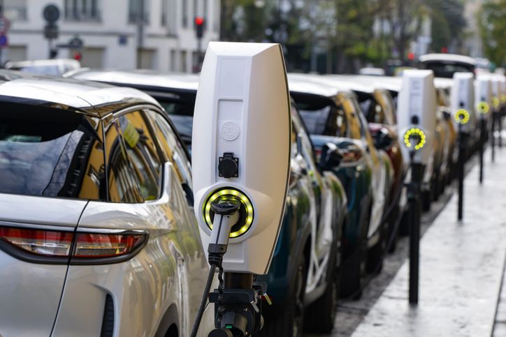 Row of electric cars charging at street-side stations, each plugged into white charging units with glowing green indicators.