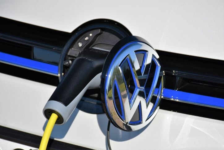 Close-up of an electric vehicle charging port with a charger plugged in, featuring a silver and blue car logo on a white background.